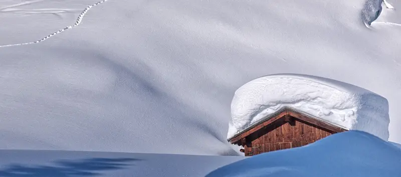 Ein Stadel unter einer meterhohen Schneedecke, dahinter blauer Himmel